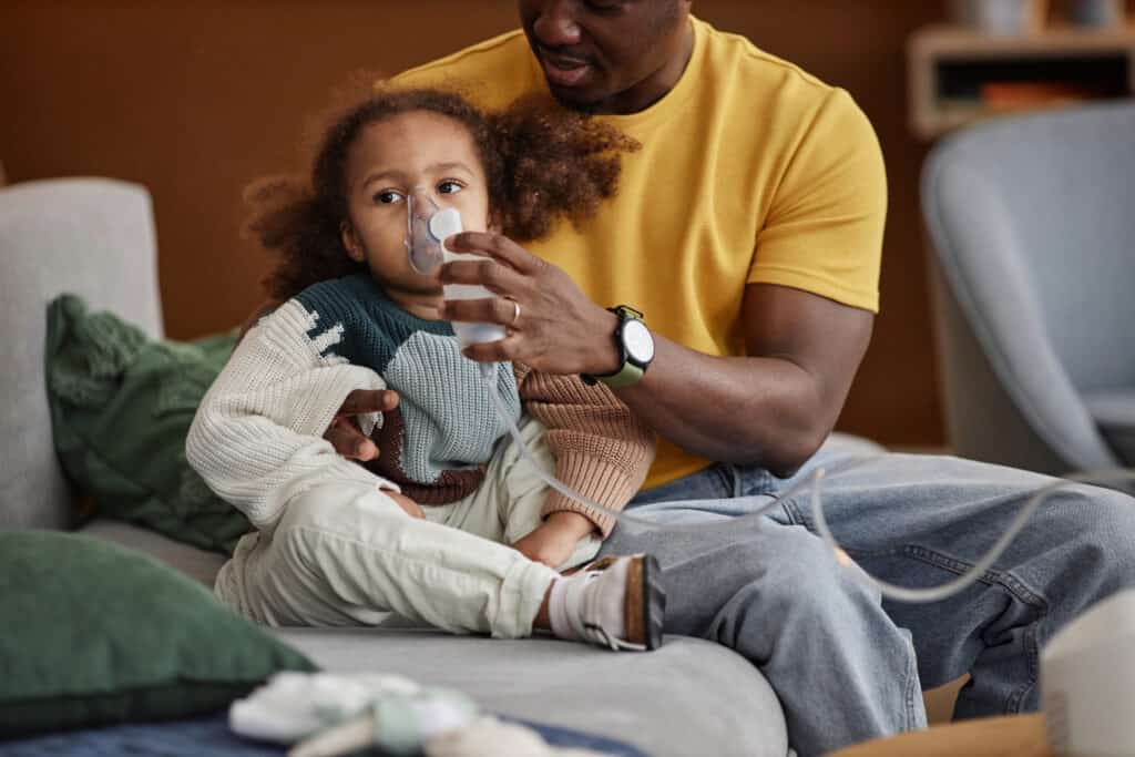 Medium long shot of African American dad embracing little daughter sitting on sofa and holding nebulizer mask on her face