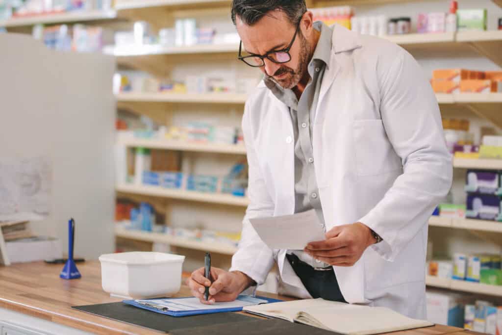 Male pharmacist writing prescription pharmacy counter. Mature druggist writing prescription on clipboard at counter.