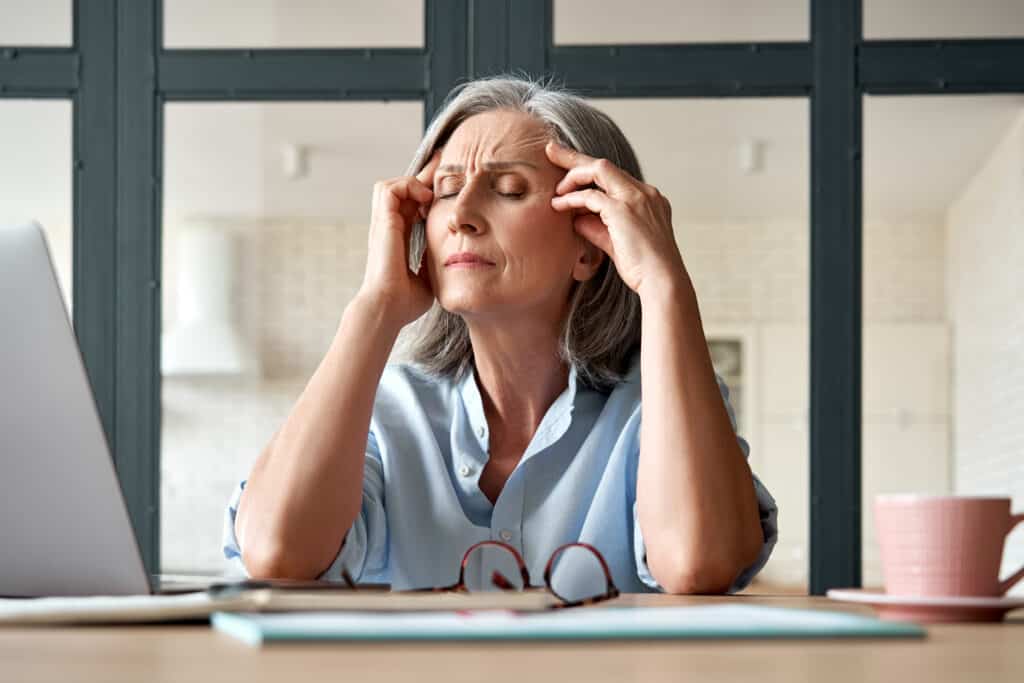 Tired stressed old mature business woman suffering from headache at work. Upset sick senior middle aged lady massaging head feeling migraine from overwork or menopause using computer at home office.