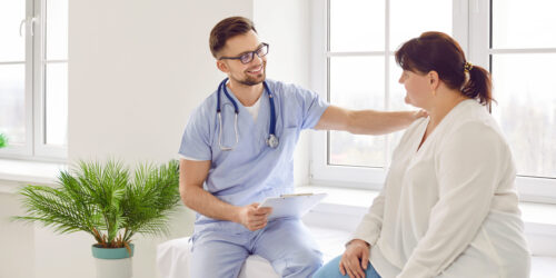 Overweight fat woman having consultation at the office. Portrait of friendly smiling doctor putting hand on shoulder supporting patient, giving consultation during medical examination in clinic.